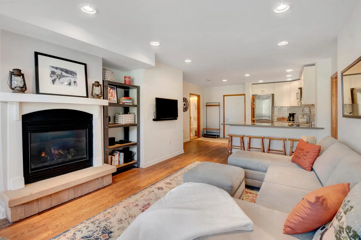 A cozy living room scene featuring a plush sofa positioned near a beautiful gas fireplace. The open-concept space includes a kitchen island with bar stools and a bookshelf, suggesting an inviting atmosphere for relaxation, conversation, and game nights. The decor and setup encourage both social gatherings and quiet moments.