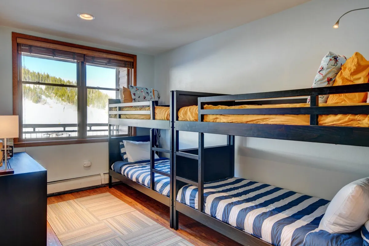 This image features a cozy room with bunk beds, a large window bringing in ample daylight, and a playful light fixture. The arrangement suggests a perfect space for families or groups, with enough beds for children or multiple guests, and a mood of warmth and togetherness.