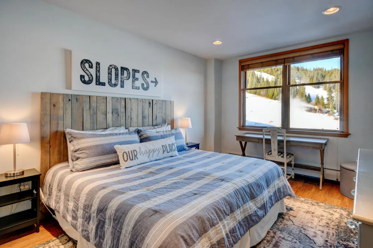 This image shows a serene bedroom with a plush bed featuring a wooden headboard, large window allowing natural light and beautiful mountain views, and decorative pillows arranged invitingly. The room's ambiance is relaxed and peaceful, emphasizing comfort and luxury.