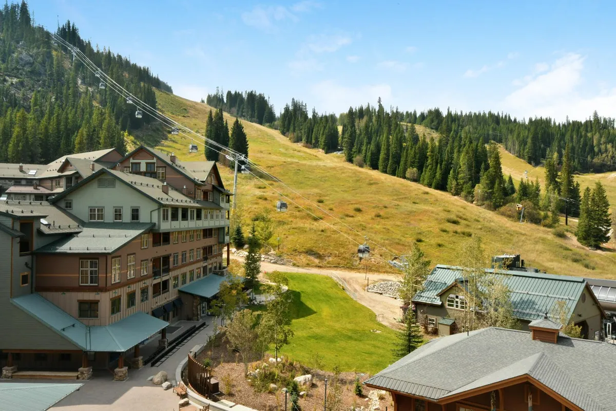 This exterior image showcases the resort buildings nestled against a dramatic mountain landscape, with a ski lift in the foreground and a green lawn hinting at summer activities. The scene captures both winter and summer possibilities in a single frame, emphasizing year-round adventure and relaxation.
