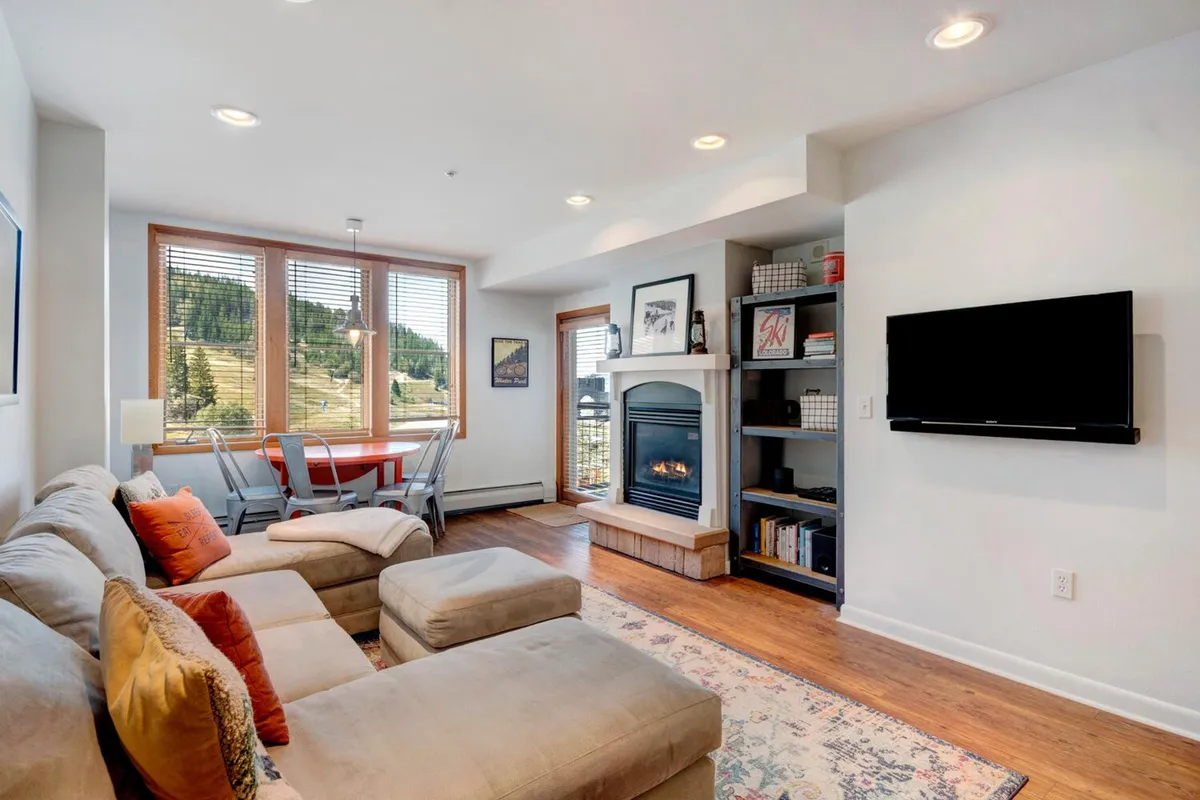 The image reveals a cozy, inviting living space featuring a crackling fireplace, expansive windows framing mountain views, a comfortable sofa, and a welcoming dining area—perfect for gathering with family or friends after a day on the slopes.