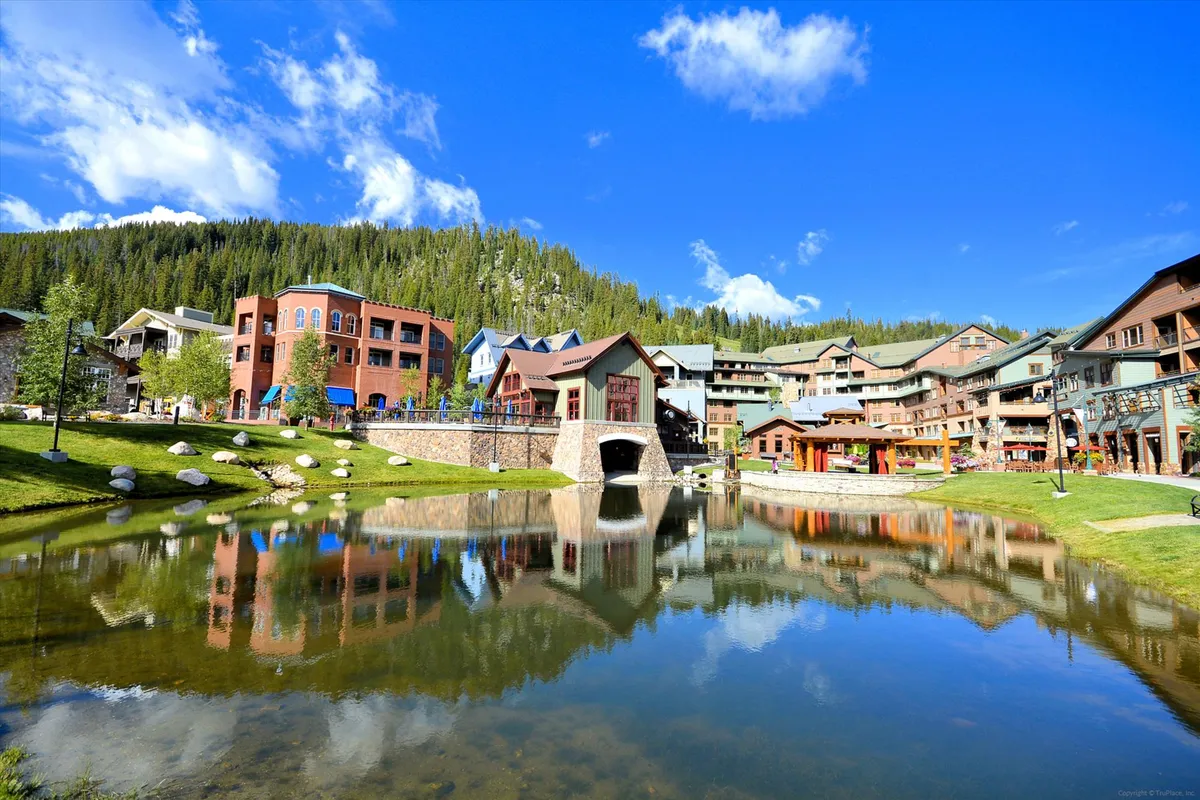 This vibrant exterior image showcases a picturesque pond bordered by a bridge and colorful buildings, framed by trees. The scene is lively and cheerful, suggesting community events, sunset reflections, and easy access to village amenities like shops and dining.