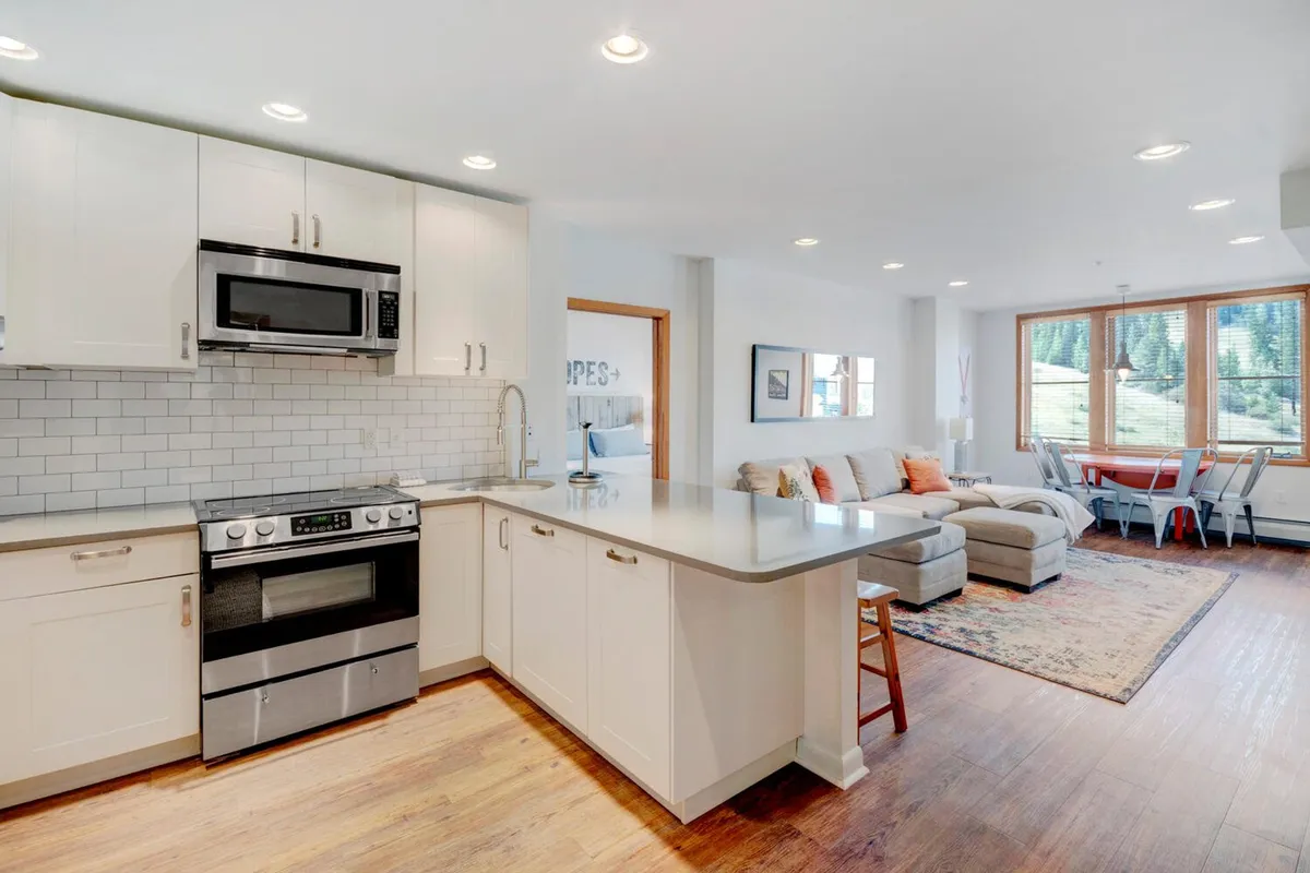 A modern kitchen scene featuring a large island, sleek appliances, comfortable seating, and expansive windows letting in natural light. The area blends cooking and relaxation, making it suitable for both meal prep and gathering with friends or family.