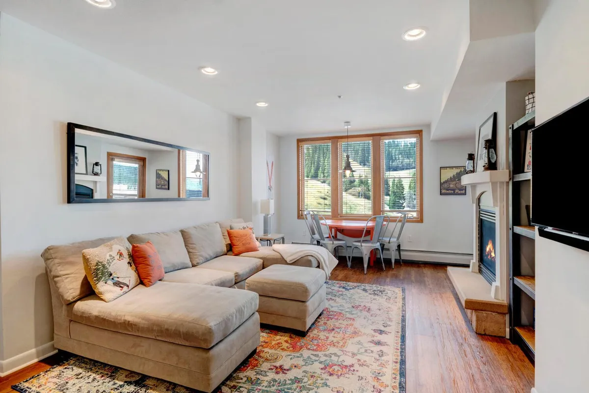 An open-plan living and dining area with a sectional sofa facing a fireplace and a dining table set next to a large window. The space feels warm and welcoming, ideal for family gatherings, meals, and playful evenings. The layout encourages easy movement between relaxing, dining, and entertaining zones.