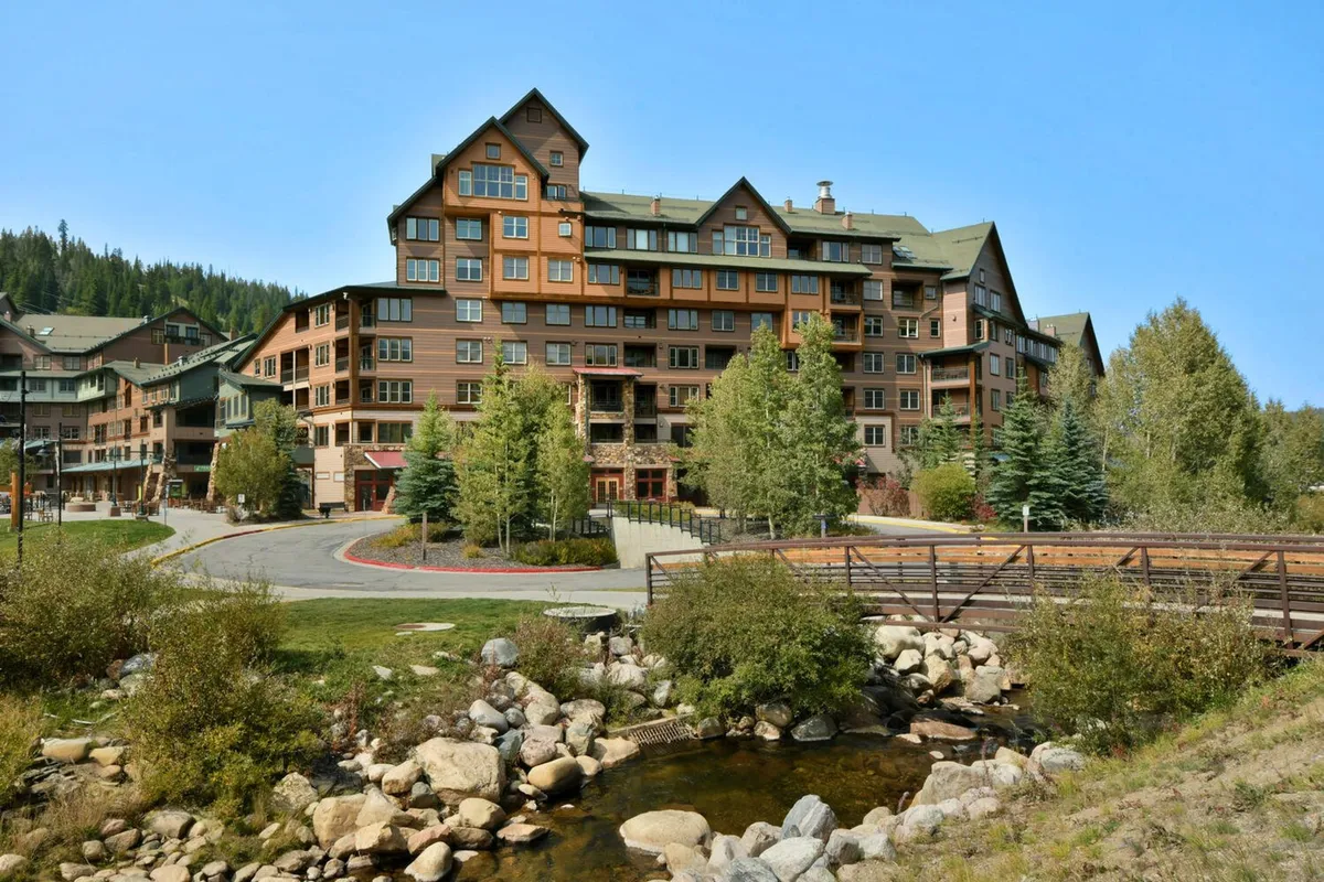 This image displays the exterior of a large, inviting building nestled among tall trees, with a tranquil stream featuring rocks and a charming pathway and bridge leading toward the property. The atmosphere is serene and welcoming, with opportunities to spot wildlife and enjoy mountain views from the building itself.