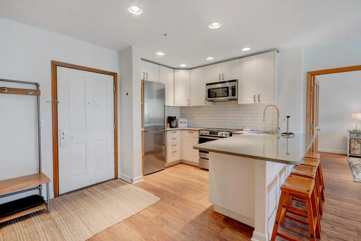 This image showcases a welcoming kitchen featuring a central island, stainless steel appliances, white cabinetry, and wooden bar stools. The space is bright and functional, ideal for both cooking and gathering. The setting conveys a sense of warmth and readiness for preparing shared meals.