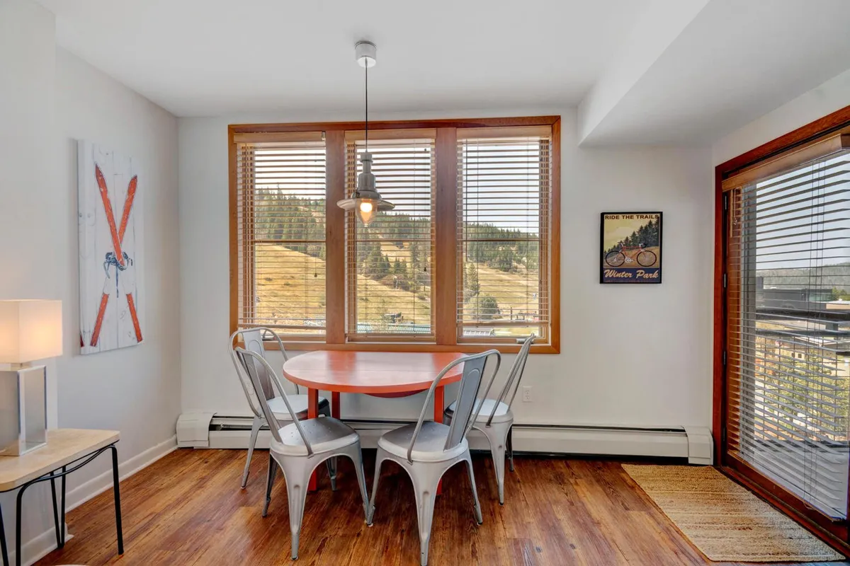 A charming dining area with a table set beside large windows framing scenic mountain views. Decorative wall art and a stylish light fixture enhance the comfortable, cheerful mood. The setting suggests enjoying meals, coffee, or board games with natural light and inspiring vistas.